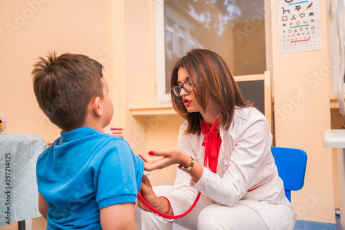 Portrait of adorable little boy visiting doctor, looking brave and smiling, holding while pediatrician listening to heartbeat with stethoscope © qunica.com