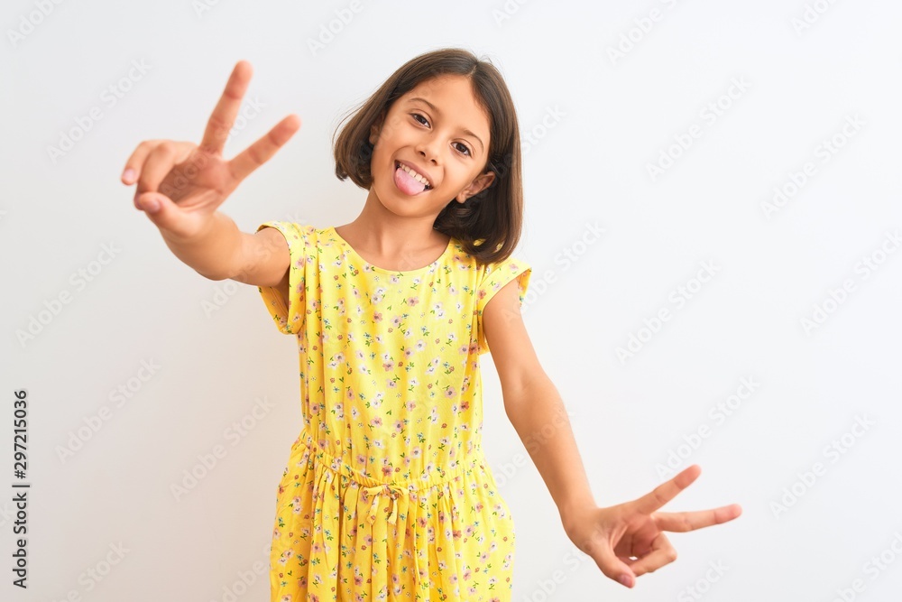 Young beautiful child girl wearing yellow floral dress standing over isolated white background smiling with tongue out showing fingers of both hands doing victory sign. Number two. Young beautiful child girl wearing yellow floral dress standing over isolated white background smiling with tongue out showing fingers of both hands doing victory sign. Number two.