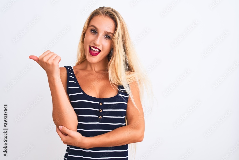 Young beautiful woman wearing casual striped t-shirt standing over isolated white background smiling with happy face looking and pointing to the side with thumb up. Young beautiful woman wearing casual striped t-shirt standing over isolated white background smiling with happy face looking and pointing to the side with thumb up.