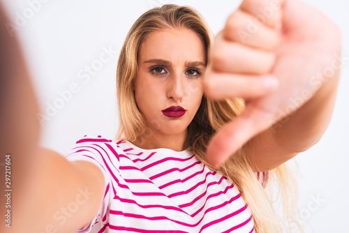 Beautiful woman wearing striped t-shirt make selfie by camera over isolated white background with angry face, negative sign showing dislike with thumbs down, rejection concept