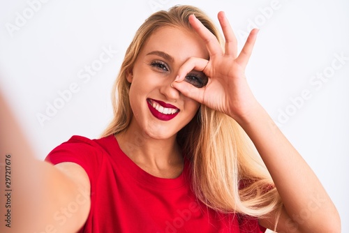 Young beautiful woman wearing t-shirt make selfie by camera over isolated white background with happy face smiling doing ok sign with hand on eye looking through fingers