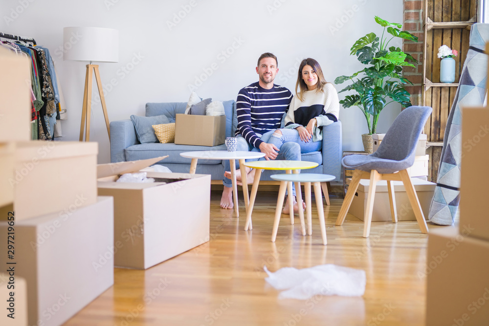 Young beautiful couple sitting on the sofa drinking cup of coffee at new home around cardboard boxes