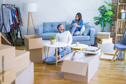 Young beautiful couple sitting on the sofa drinking cup of coffee at new home around cardboard boxes