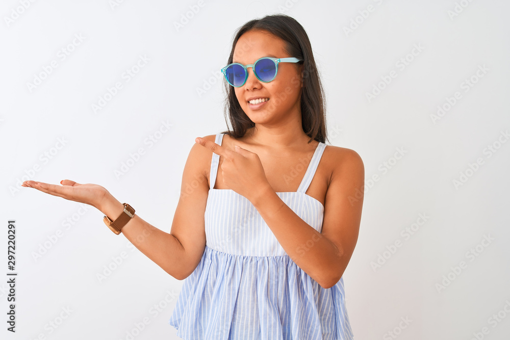 Young chinese woman wearing striped dress and sunglasses over isolated white background amazed and smiling to the camera while presenting with hand and pointing with finger. Young chinese woman wearing striped dress and sunglasses over isolated white background amazed and smiling to the camera while presenting with hand and pointing with finger.