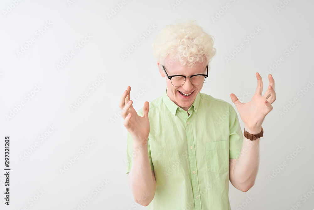 Young albino blond man wearing green shirt and glasses over isolated white background celebrating mad and crazy for success with arms raised and closed eyes screaming excited. Winner concept Young albino blond man wearing green shirt and glasses over isolated white background celebrating mad and crazy for success with arms raised and closed eyes screaming excited. Winner concept