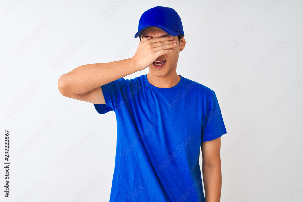 Chinese deliveryman wearing blue t-shirt and cap standing over isolated white background smiling and laughing with hand on face covering eyes for surprise. Blind concept.