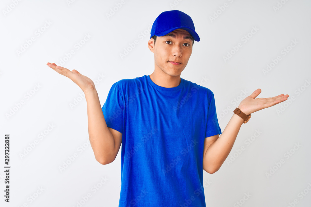 Chinese deliveryman wearing blue t-shirt and cap standing over isolated white background clueless and confused expression with arms and hands raised. Doubt concept.