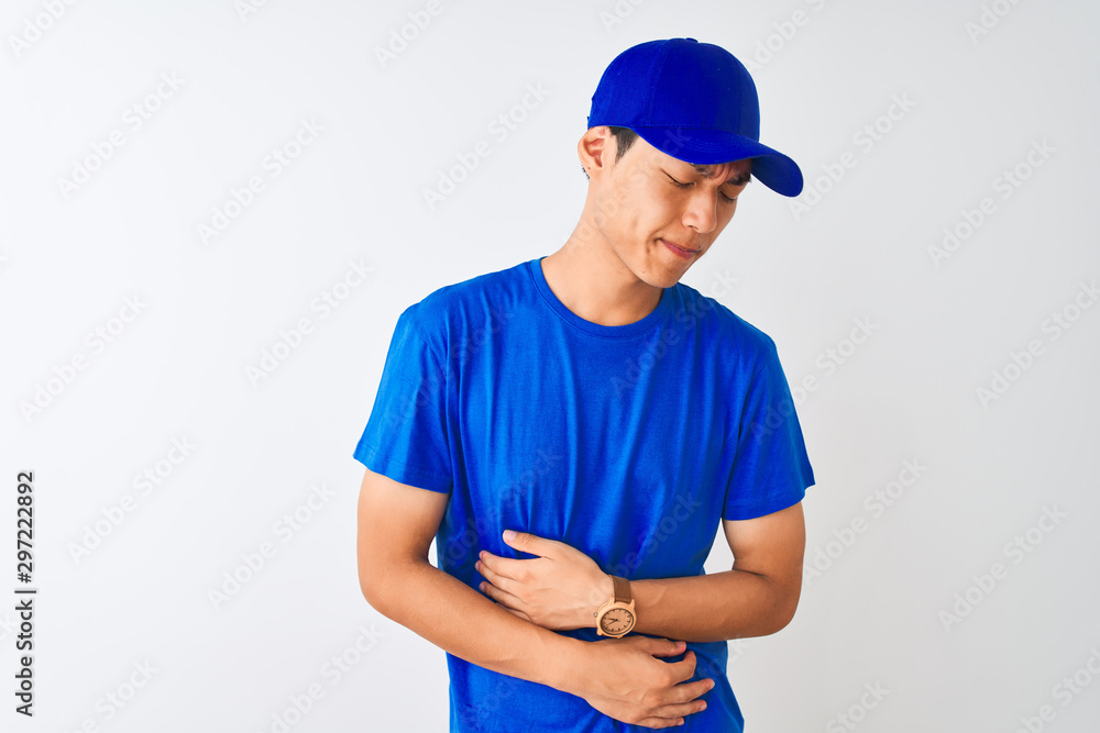 Chinese deliveryman wearing blue t-shirt and cap standing over isolated white background with hand on stomach because nausea, painful disease feeling unwell. Ache concept.