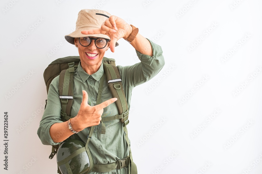 Middle age hiker woman wearing backpack canteen hat glasses over isolated white background smiling making frame with hands and fingers with happy face. Creativity and photography concept.