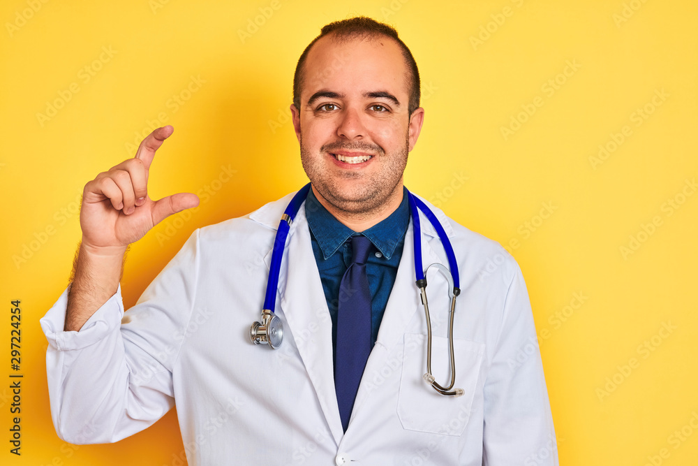 Young doctor man wearing coat and stethoscope standing over isolated yellow background smiling and confident gesturing with hand doing small size sign with fingers looking and the camera. Measure Young doctor man wearing coat and stethoscope standing over isolated yellow background smiling and confident gesturing with hand doing small size sign with fingers looking and the camera. Measure