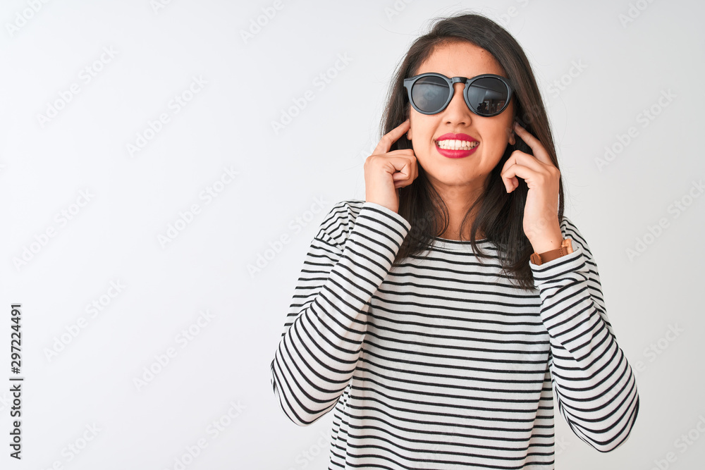 Chinese woman wearing striped t-shirt and sunglasses standing over isolated white background covering ears with fingers with annoyed expression for the noise of loud music. Deaf concept. Chinese woman wearing striped t-shirt and sunglasses standing over isolated white background covering ears with fingers with annoyed expression for the noise of loud music. Deaf concept.