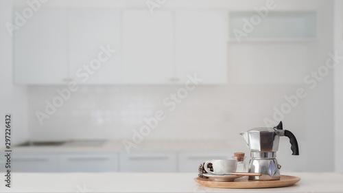 Coffee cup and Moka pot in wooden tray on white counter with blurred kitchen background photo