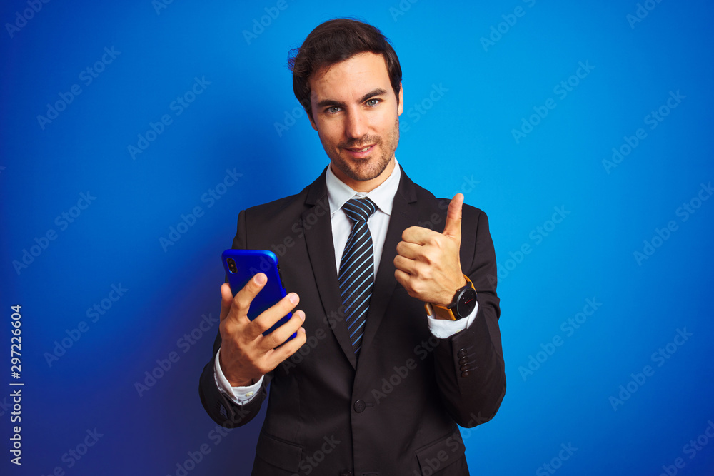 Young handsome businessman using smartphone standing over isolated blue background happy with big smile doing ok sign, thumb up with fingers, excellent sign Young handsome businessman using smartphone standing over isolated blue background happy with big smile doing ok sign, thumb up with fingers, excellent sign