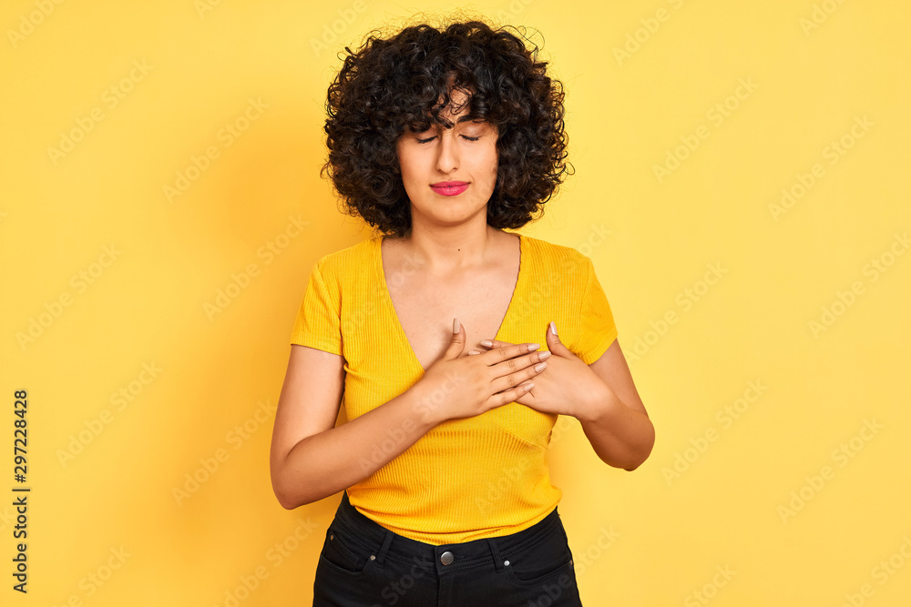 Young arab woman with curly hair wearing t-shirt standing over isolated yellow background smiling with hands on chest with closed eyes and grateful gesture on face. Health concept. Young arab woman with curly hair wearing t-shirt standing over isolated yellow background smiling with hands on chest with closed eyes and grateful gesture on face. Health concept.