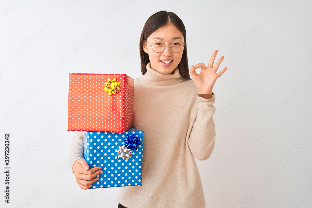 Young chinese woman holding birthday gifts over isolated white background smiling positive doing ok sign with hand and fingers. Successful expression.