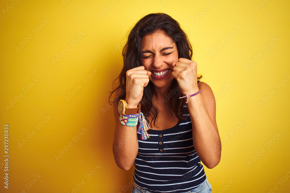 Young beautiful woman wearing striped t-shirt standing over isolated yellow background very happy and excited doing winner gesture with arms raised, smiling and screaming for success Young beautiful woman wearing striped t-shirt standing over isolated yellow background very happy and excited doing winner gesture with arms raised, smiling and screaming for success