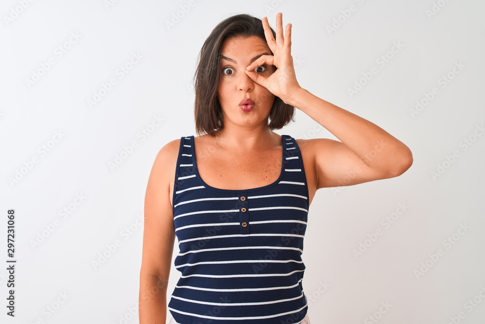 Young beautiful woman wearing blue striped t-shirt standing over isolated white background doing ok gesture shocked with surprised face, eye looking through fingers. Unbelieving expression. Young beautiful woman wearing blue striped t-shirt standing over isolated white background doing ok gesture shocked with surprised face, eye looking through fingers. Unbelieving expression.