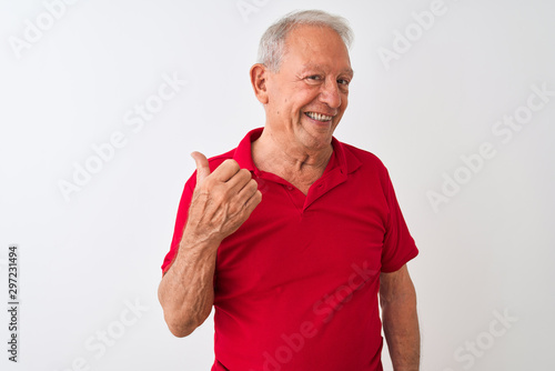 Senior grey-haired man wearing red polo standing over isolated white background smiling with happy face looking and pointing to the side with thumb up.