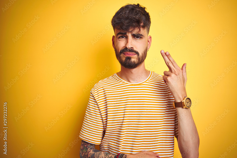 Young man with tattoo wearing striped t-shirt standing over isolated yellow background Shooting and killing oneself pointing hand and fingers to head like gun, suicide gesture. Young man with tattoo wearing striped t-shirt standing over isolated yellow background Shooting and killing oneself pointing hand and fingers to head like gun, suicide gesture.