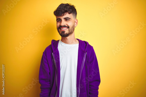 Young man with tattoo wearing sport purple sweatshirt over isolated yellow background looking away to side with smile on face, natural expression. Laughing confident.