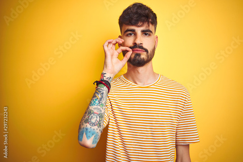 Young man with tattoo wearing striped t-shirt standing over isolated yellow background mouth and lips shut as zip with fingers. Secret and silent, taboo talking