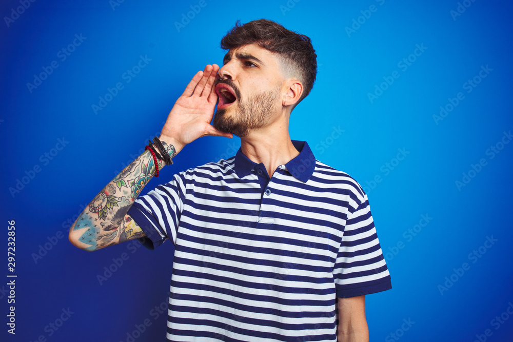 Young man with tattoo wearing striped polo standing over isolated blue background shouting and screaming loud to side with hand on mouth. Communication concept.
