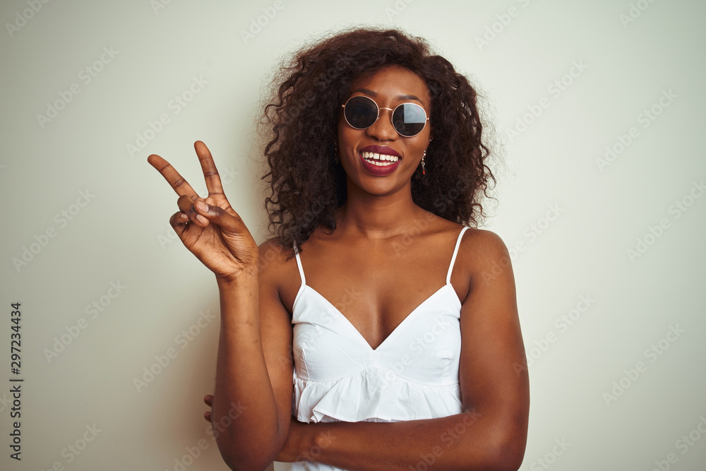 Young african american woman wearing t-shirt and sunglasses over isolated white background smiling with happy face winking at the camera doing victory sign. Number two.