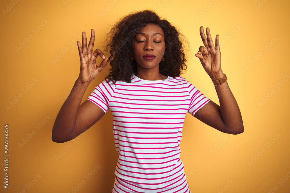 Young african american woman wearing striped t-shirt over isolated yellow background relax and smiling with eyes closed doing meditation gesture with fingers. Yoga concept. Young african american woman wearing striped t-shirt over isolated yellow background relax and smiling with eyes closed doing meditation gesture with fingers. Yoga concept.