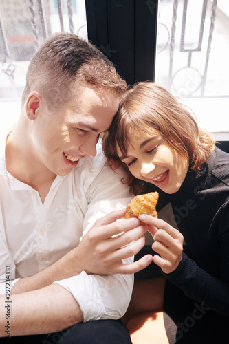 Smiling young man feeding pretty girlfriend with small croissant with sesame seeds