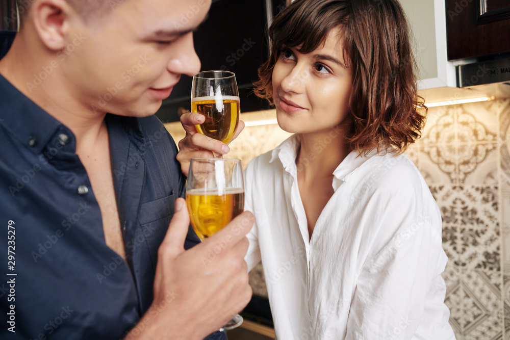 Beautiful young woman looking at pensive boyfriend when they are drinking tasty white wine in kitchen Beautiful young woman looking at pensive boyfriend when they are drinking tasty white wine in kitchen