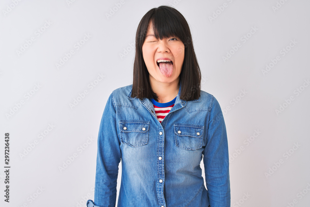 Young beautiful chinese woman wearing denim shirt over isolated white background sticking tongue out happy with funny expression. Emotion concept. Young beautiful chinese woman wearing denim shirt over isolated white background sticking tongue out happy with funny expression. Emotion concept.