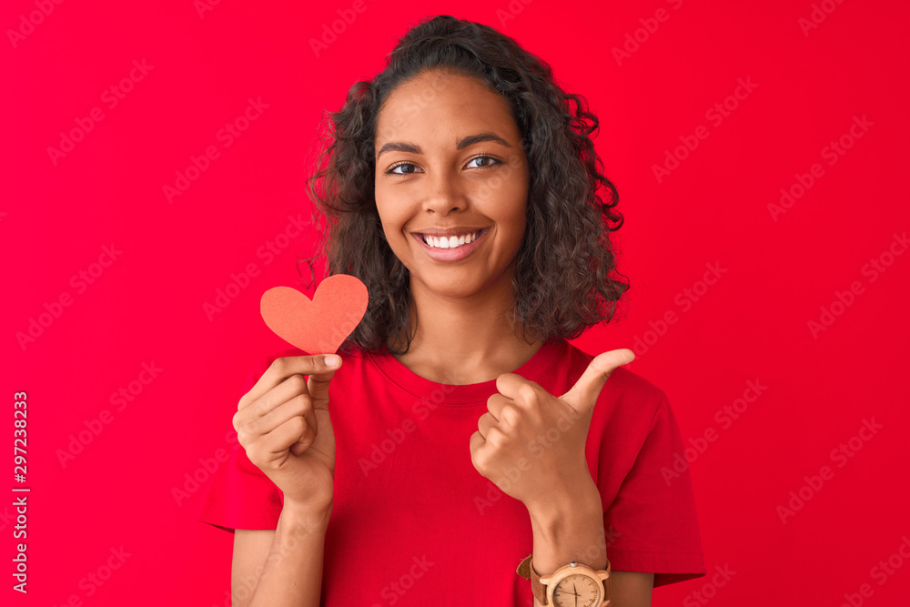 Young brazilian woman holding paper heart standing over isolated red background happy with big smile doing ok sign, thumb up with fingers, excellent sign Young brazilian woman holding paper heart standing over isolated red background happy with big smile doing ok sign, thumb up with fingers, excellent sign