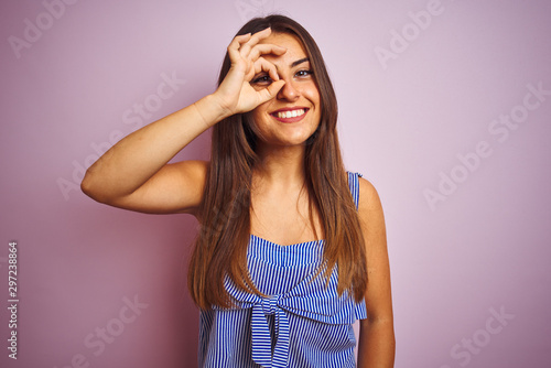 Young beautiful woman wearing striped dress standing over isolated pink background doing ok gesture with hand smiling, eye looking through fingers with happy face.