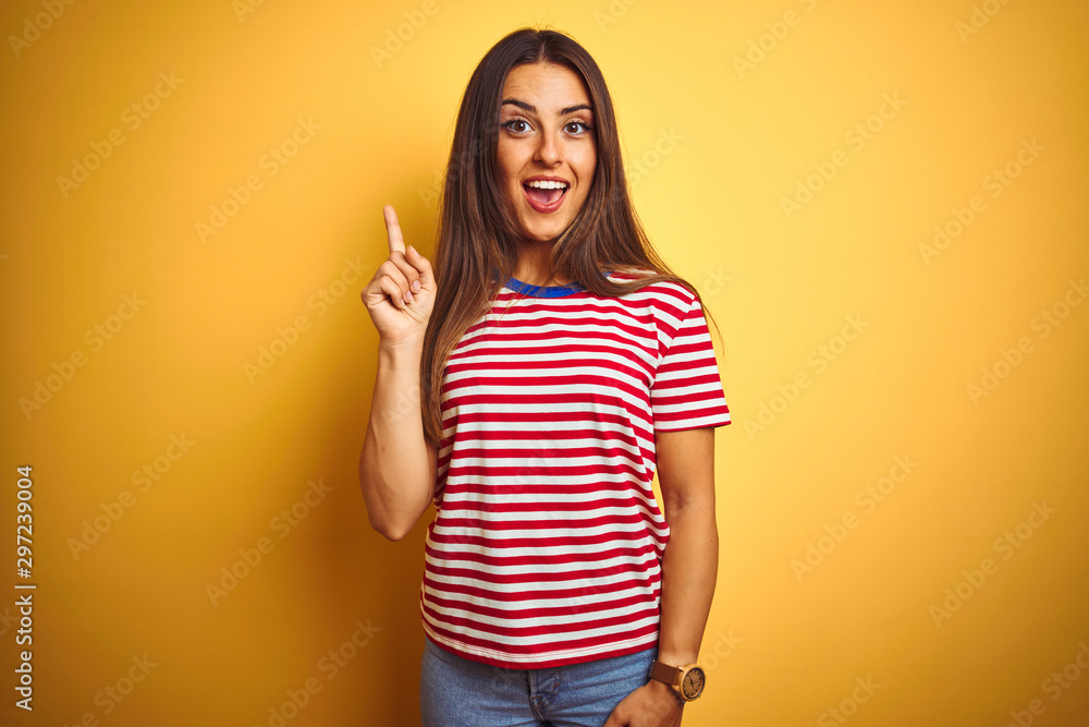 Young beautiful woman wearing striped t-shirt standing over isolated yellow background pointing finger up with successful idea. Exited and happy. Number one. Young beautiful woman wearing striped t-shirt standing over isolated yellow background pointing finger up with successful idea. Exited and happy. Number one.