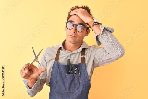 Handsome hairdresser man wearing apron holding scissors over isolated yellow background stressed with hand on head, shocked with shame and surprise face, angry and frustrated. Fear 