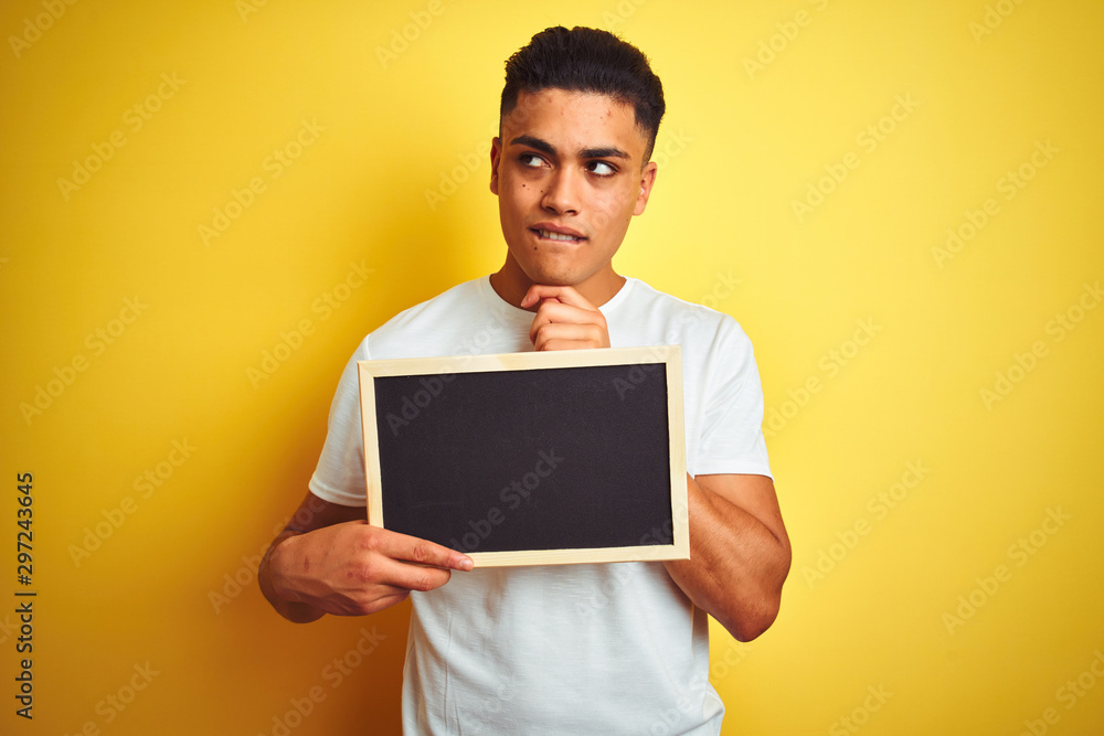 Young brazilian man holding blackboard standing over isolated yellow background serious face thinking about question, very confused idea Young brazilian man holding blackboard standing over isolated yellow background serious face thinking about question, very confused idea