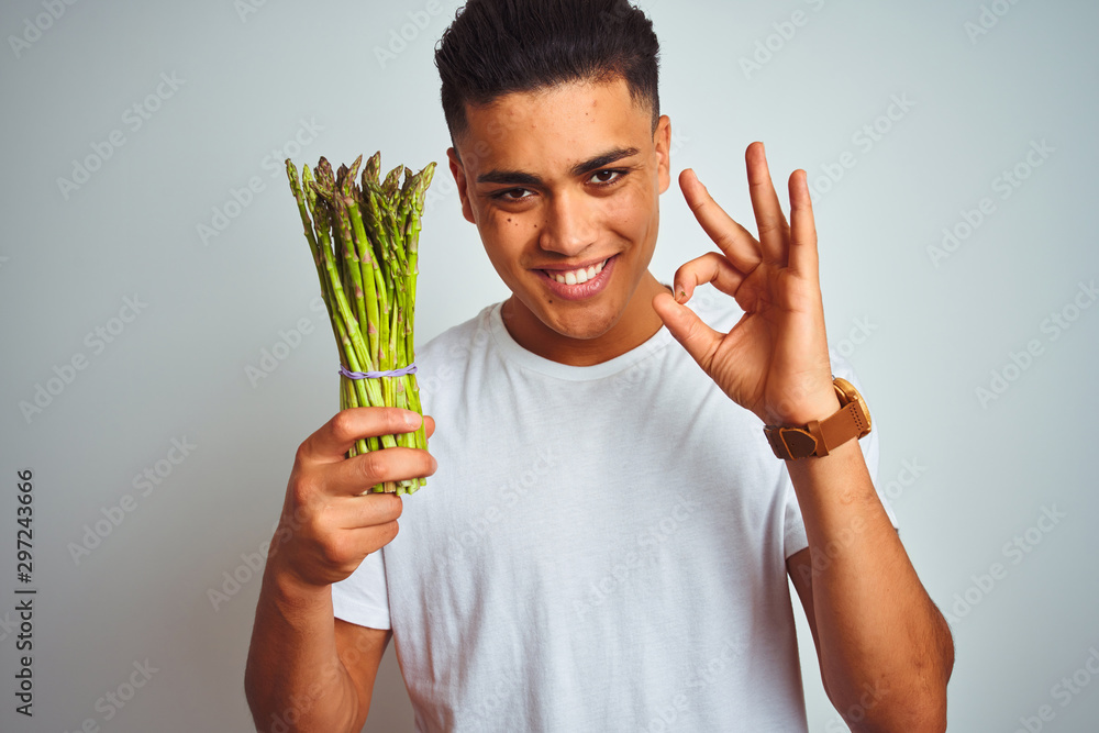 Young brazilian man eating asparagus standing over isolated grey background doing ok sign with fingers, excellent symbol Young brazilian man eating asparagus standing over isolated grey background doing ok sign with fingers, excellent symbol