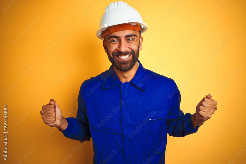 Handsome indian worker man wearing uniform and helmet over isolated yellow background celebrating surprised and amazed for success with arms raised and open eyes. Winner concept. Handsome indian worker man wearing uniform and helmet over isolated yellow background celebrating surprised and amazed for success with arms raised and open eyes. Winner concept.