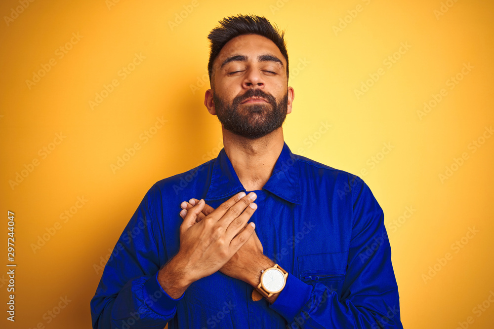 Handsome indian worker man wearing uniform over isolated yellow background smiling with hands on chest with closed eyes and grateful gesture on face. Health concept. Handsome indian worker man wearing uniform over isolated yellow background smiling with hands on chest with closed eyes and grateful gesture on face. Health concept.