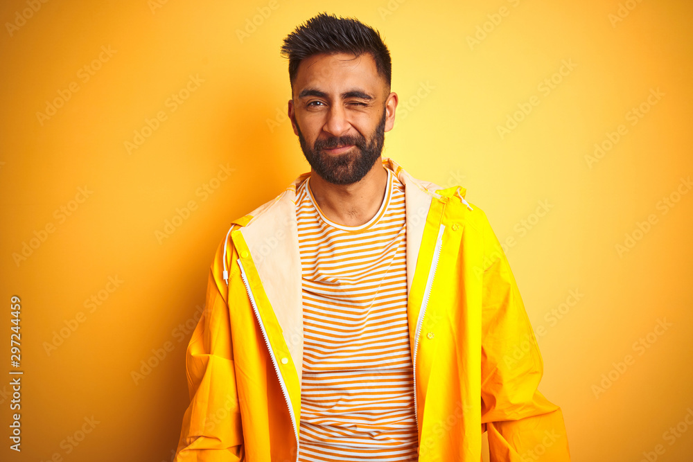 Young indian man wearing raincoat standing over isolated yellow background winking looking at the camera with sexy expression, cheerful and happy face. Young indian man wearing raincoat standing over isolated yellow background winking looking at the camera with sexy expression, cheerful and happy face.
