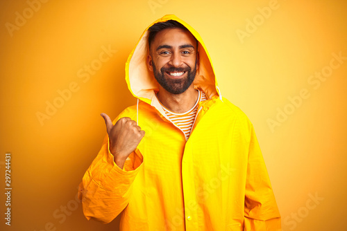 Young indian man wearing raincoat with hood standing over isolated yellow background smiling with happy face looking and pointing to the side with thumb up. © Krakenimages.com
