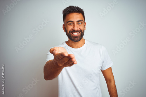 Young indian man wearing t-shirt standing over isolated white background smiling cheerful offering palm hand giving assistance and acceptance.