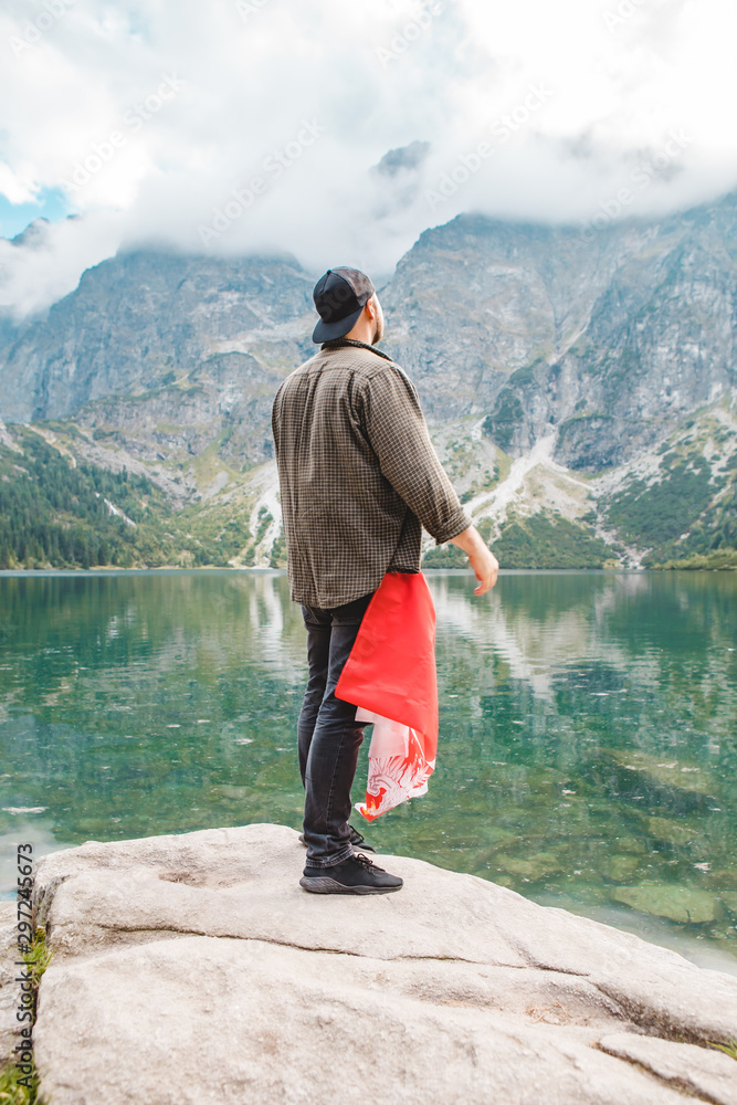 man standing at the edge looking at lake in tatra mountains with poland flag in pocket