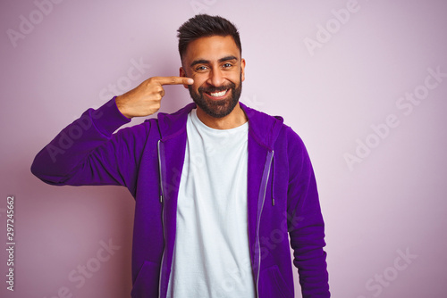 Young indian man wearing purple sweatshirt standing over isolated pink background Pointing with hand finger to face and nose, smiling cheerful. Beauty concept © Krakenimages.com