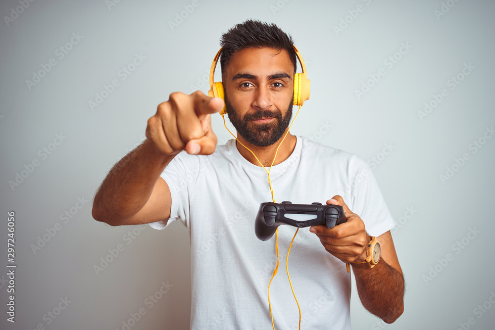 Arab indian gamer man playing video game using headphones over isolated white background pointing with finger to the camera and to you, hand sign, positive and confident gesture from the front Arab indian gamer man playing video game using headphones over isolated white background pointing with finger to the camera and to you, hand sign, positive and confident gesture from the front