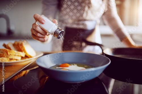 Close up of caucasian woman adding salt in sunny side up eggs while standing in kitchen next to stove.