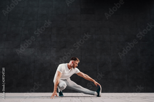 Full length of attractive Caucasian bearded blonde man stretching leg. In background is gray wall.