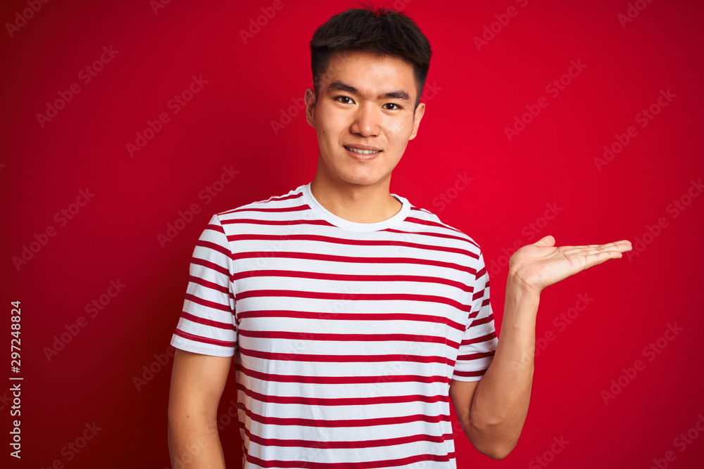 Young asian chinese man wearing striped t-shirt standing over isolated red background smiling cheerful presenting and pointing with palm of hand looking at the camera. Young asian chinese man wearing striped t-shirt standing over isolated red background smiling cheerful presenting and pointing with palm of hand looking at the camera.
