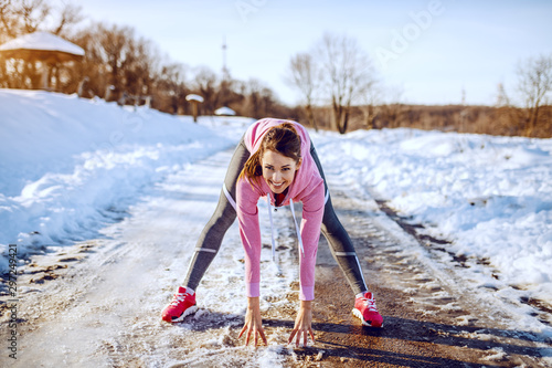 Cheerful beautiful caucasian brunette in sportswear and with ponytail doing warm up exercises while bending forward and looking at camera. Outdoor fitness. Winter time. photo