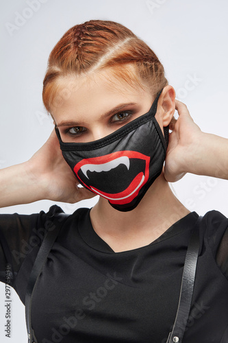 Close-up portrait of a ginger girl with three french braids, posing on a grey background. She's wearing black sweatshirt and leather belt. Her face is hidden by black mask with vampire mouth print.  photo